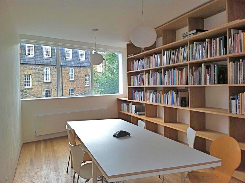 Foto på ett mellanstort funkis hemmabibliotek, med vita väggar, mörkt trägolv och ett fristående skrivbord