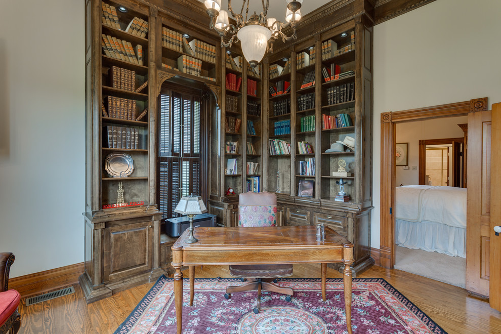 Cette image montre un bureau victorien de taille moyenne avec une bibliothèque ou un coin lecture, un mur blanc, un sol en bois brun, un bureau indépendant et un sol marron.