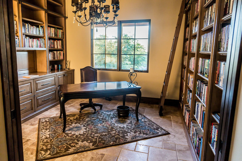 Lantlig inredning av ett mycket stort arbetsrum, med ett bibliotek, beige väggar, travertin golv, en standard öppen spis, en spiselkrans i sten, ett fristående skrivbord och flerfärgat golv