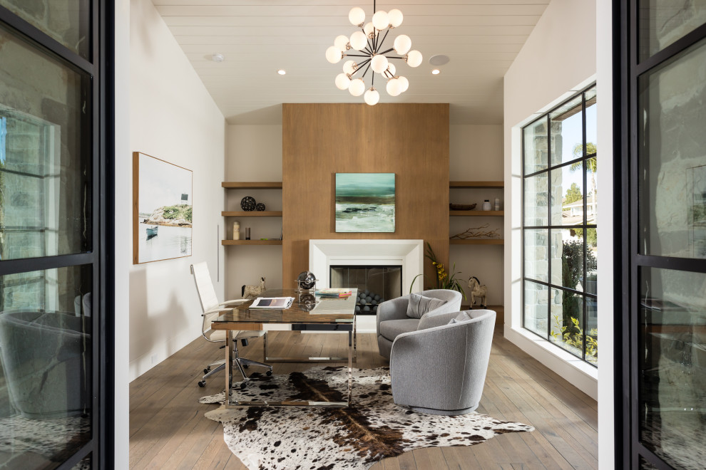 Landhaus Arbeitszimmer mit Arbeitsplatz, weißer Wandfarbe, braunem Holzboden, Kamin, Kaminumrandung aus Holz, freistehendem Schreibtisch und braunem Boden in Orange County