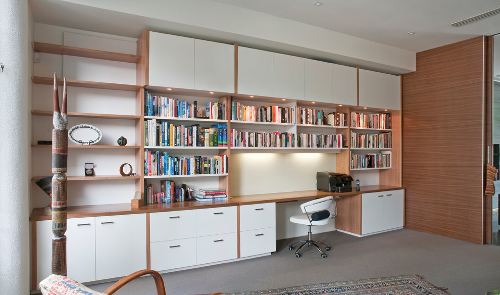 Modern inredning av ett stort hemmabibliotek, med vita väggar, heltäckningsmatta och ett inbyggt skrivbord