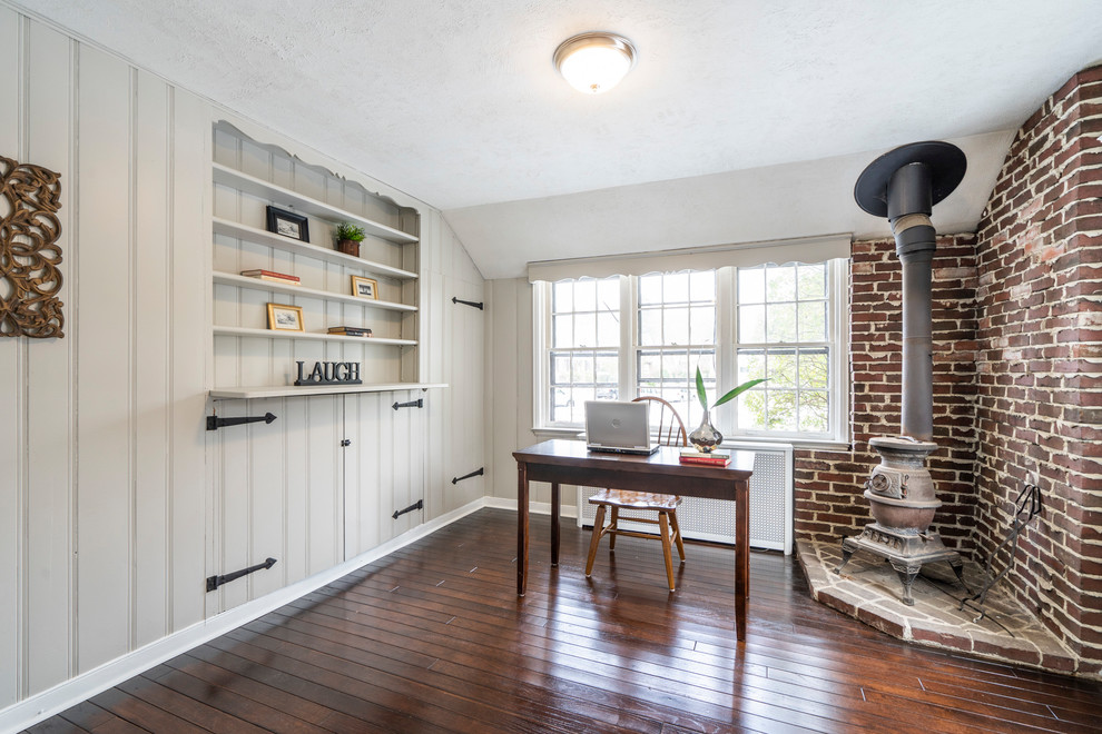 Ejemplo de despacho rural con paredes grises, estufa de leña, marco de chimenea de ladrillo, escritorio independiente y suelo marrón
