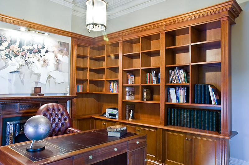 Klassisk inredning av ett stort hemmabibliotek, med beige väggar, mellanmörkt trägolv, en standard öppen spis, en spiselkrans i trä och ett fristående skrivbord