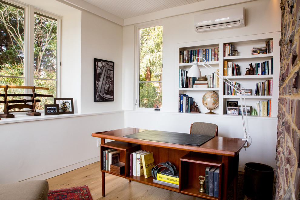 Bild på ett mellanstort funkis hemmabibliotek, med vita väggar, mellanmörkt trägolv, ett fristående skrivbord och beiget golv