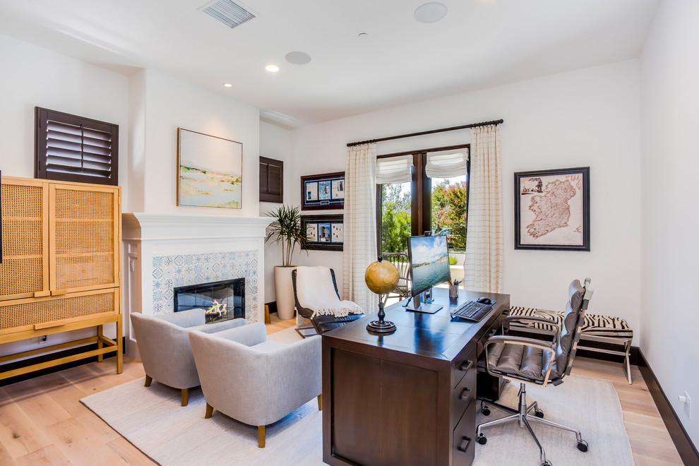 Inspiration for a mediterranean study in Orange County with white walls, a standard fireplace, a tiled fireplace surround, a freestanding desk, light hardwood flooring and beige floors.