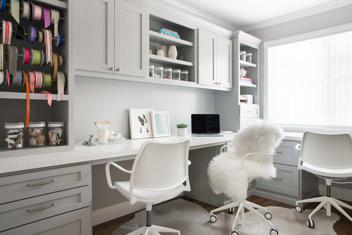 Premium RTA cabinets Canada: light gray shaker built-ins in a bright craft room/home office with a wraparound desk, open shelves, ribbon organizer, jars, and white swivel chairs by a large window.