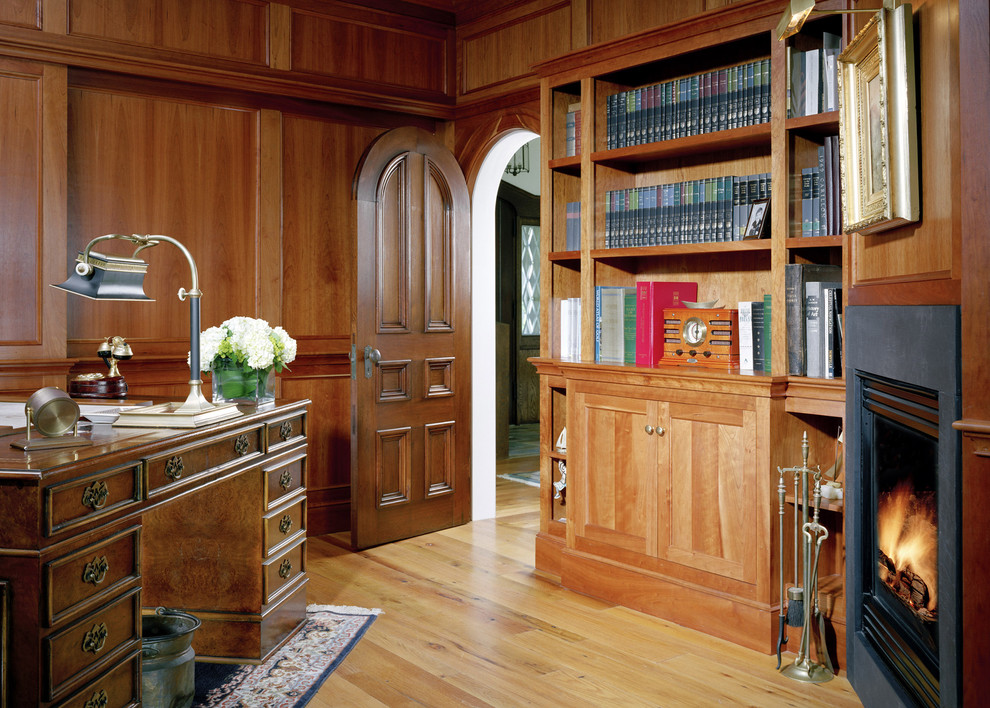 Klassisk inredning av ett mellanstort hemmabibliotek, med bruna väggar, ljust trägolv, en standard öppen spis, ett fristående skrivbord och brunt golv