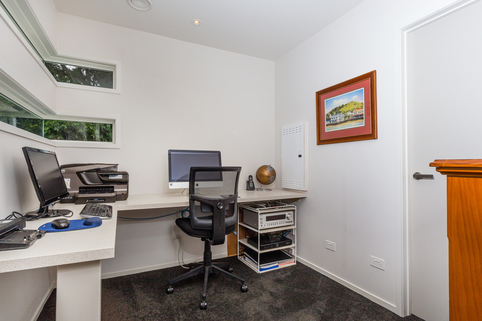 Bild på ett litet arbetsrum, med vita väggar, heltäckningsmatta, ett inbyggt skrivbord och grått golv