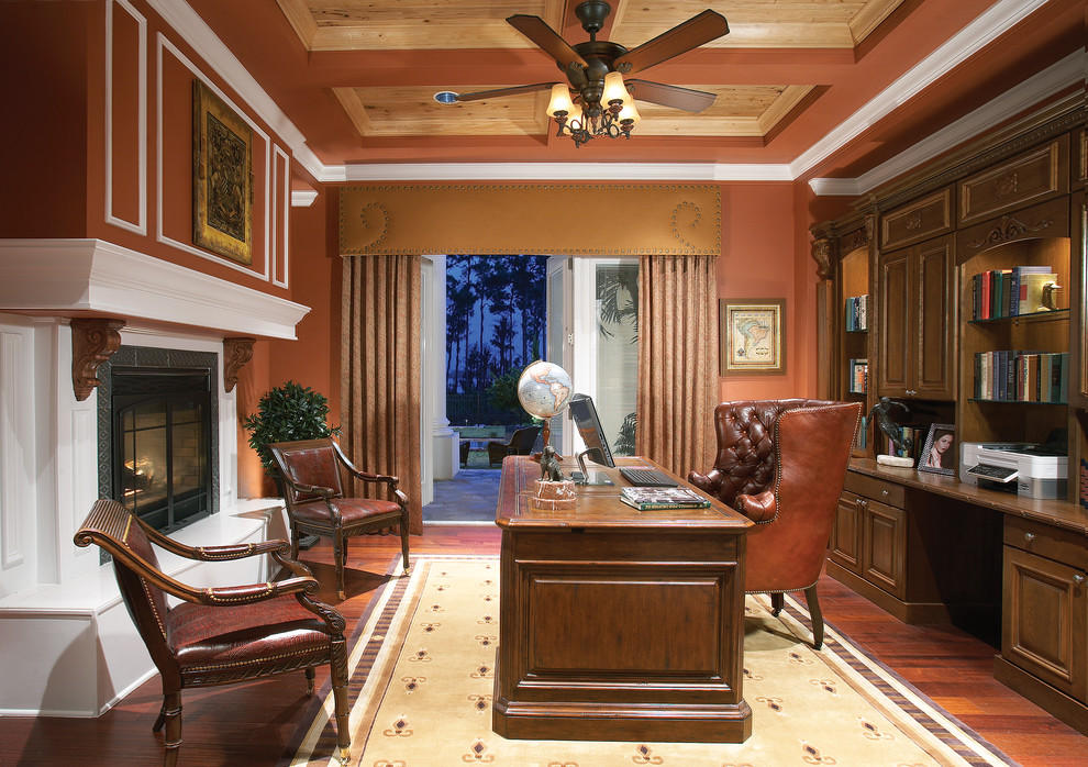 На фото: кабинет в классическом стиле с красными стенами, темным паркетным полом, стандартным камином и отдельно стоящим рабочим столом