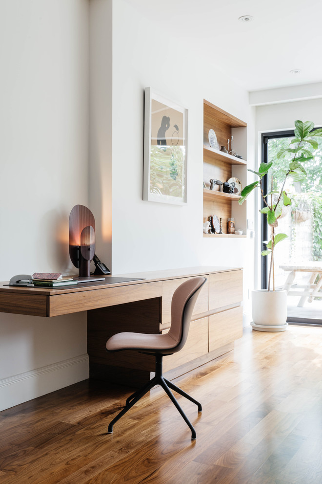 Mittelgroßes Modernes Arbeitszimmer mit weißer Wandfarbe, braunem Holzboden, Einbau-Schreibtisch und braunem Boden in New York