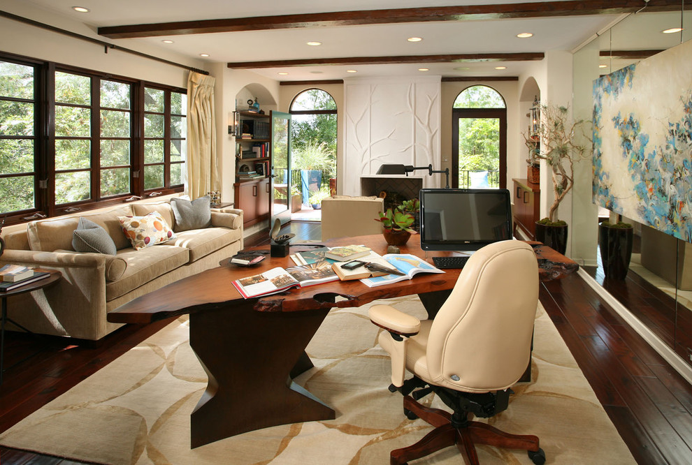 This is an example of a large classic study in Los Angeles with dark hardwood flooring, a freestanding desk, beige walls, a standard fireplace, a plastered fireplace surround and brown floors.