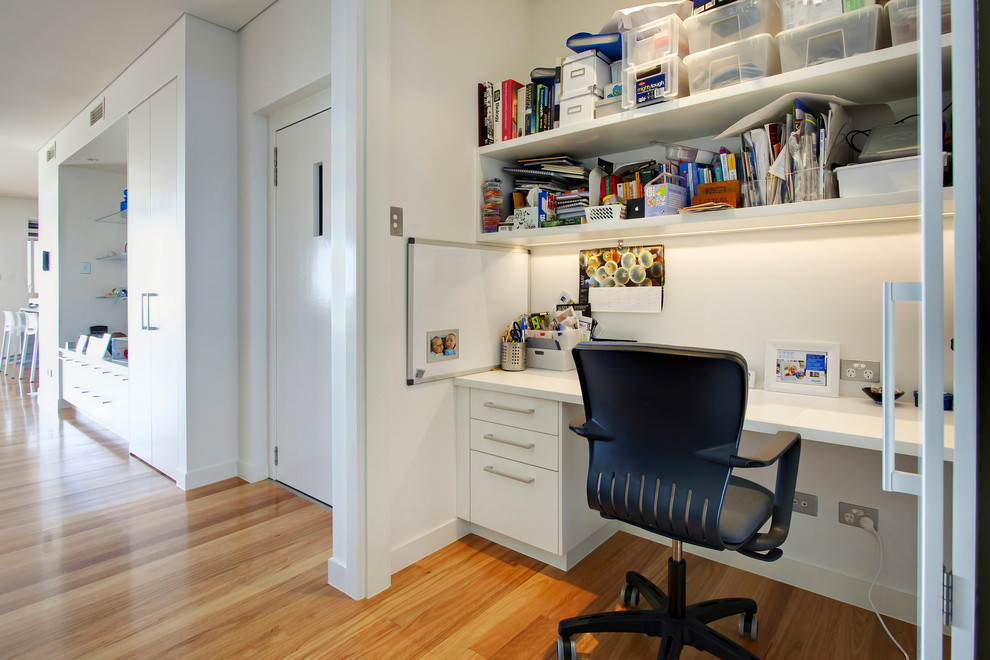 Ejemplo de despacho actual pequeño sin chimenea con paredes blancas, escritorio empotrado, suelo de madera clara y suelo marrón