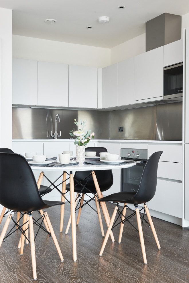 Eat-in kitchen - small contemporary l-shaped dark wood floor and brown floor eat-in kitchen idea in London with an undermount sink, flat-panel cabinets, white cabinets, solid surface countertops, metallic backsplash, metal backsplash, black appliances and white countertops