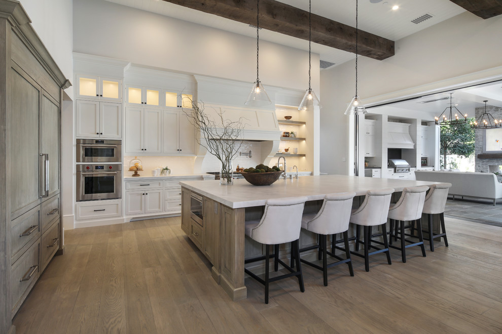 Immagine di una cucina chic con ante in stile shaker, ante bianche, paraspruzzi bianco, elettrodomestici in acciaio inossidabile, pavimento in legno massello medio, pavimento marrone e top bianco