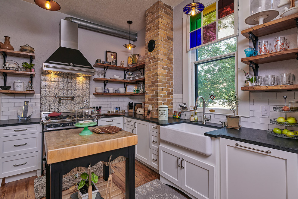Example of a mid-sized eclectic l-shaped medium tone wood floor and brown floor eat-in kitchen design in Other with a farmhouse sink, shaker cabinets, white cabinets, granite countertops, white backsplash, porcelain backsplash, stainless steel appliances, an island and black countertops