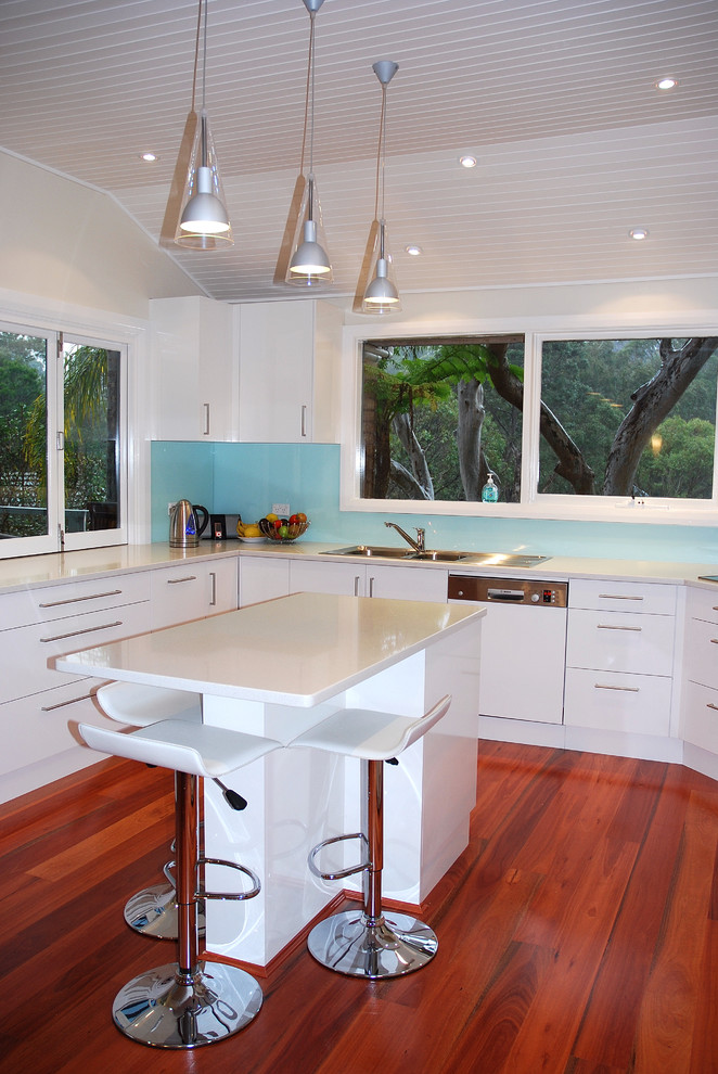 Moderne Küche in U-Form mit Doppelwaschbecken, flächenbündigen Schrankfronten, weißen Schränken, Küchenrückwand in Blau, Glasrückwand, weißen Elektrogeräten, braunem Holzboden und Kücheninsel in Sydney