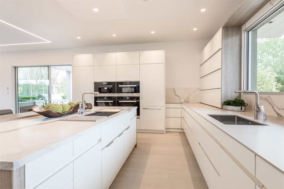 Inspiration for an expansive contemporary u-shaped kitchen/diner in New York with a submerged sink, flat-panel cabinets, white cabinets, white splashback, marble splashback, stainless steel appliances, light hardwood flooring, an island and beige floors.