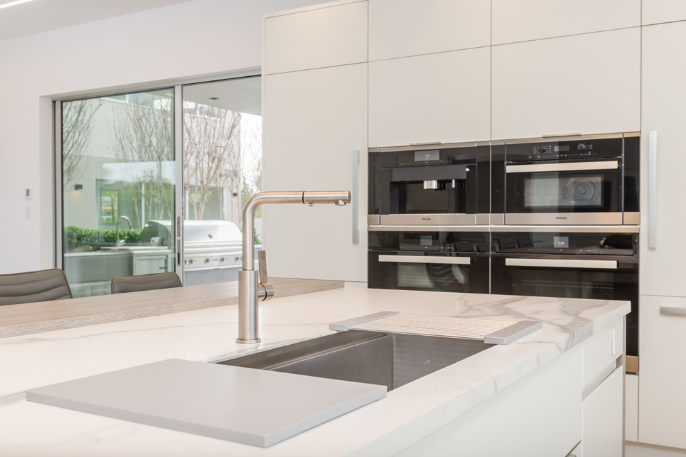 This is an example of an expansive contemporary u-shaped kitchen/diner in New York with a submerged sink, flat-panel cabinets, white cabinets, white splashback, marble splashback, stainless steel appliances, light hardwood flooring, an island and beige floors.