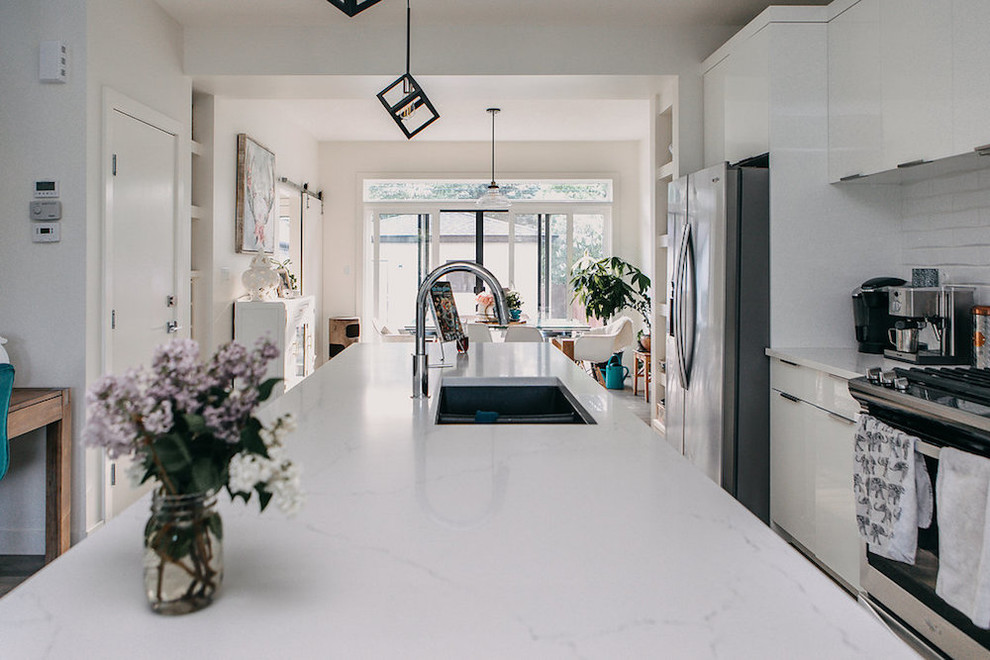Inredning av ett modernt mellanstort vit linjärt vitt kök med öppen planlösning, med en undermonterad diskho, skåp i shakerstil, vita skåp, vitt stänkskydd, stänkskydd i tunnelbanekakel, rostfria vitvaror, ljust trägolv, en köksö och brunt golv