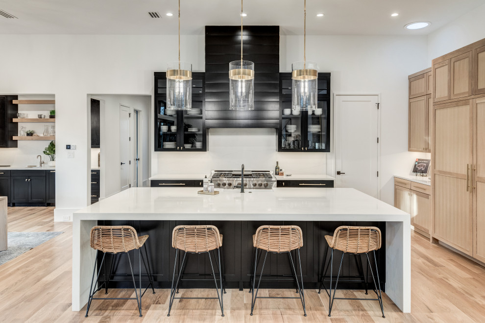 Photo of a modern l-shaped open plan kitchen in Dallas with a belfast sink, recessed-panel cabinets, black cabinets, engineered stone countertops, white splashback, marble splashback, stainless steel appliances, light hardwood flooring, an island, brown floors and white worktops.