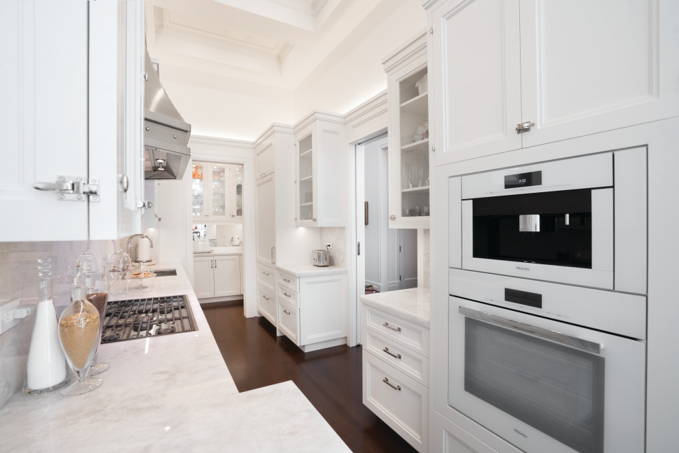 Example of a transitional u-shaped dark wood floor, brown floor and coffered ceiling enclosed kitchen design in New York with an undermount sink, recessed-panel cabinets, white cabinets, marble countertops, gray backsplash, marble backsplash, white appliances and no island
