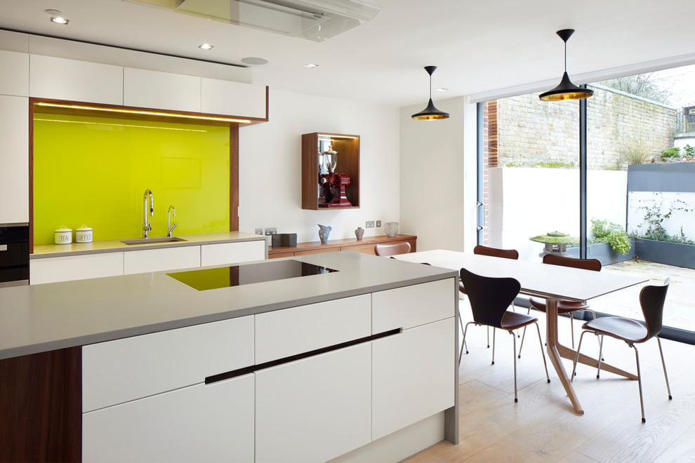 Design ideas for a contemporary kitchen/diner in London with flat-panel cabinets, white cabinets, green splashback, glass sheet splashback, light hardwood flooring, an island and a submerged sink.