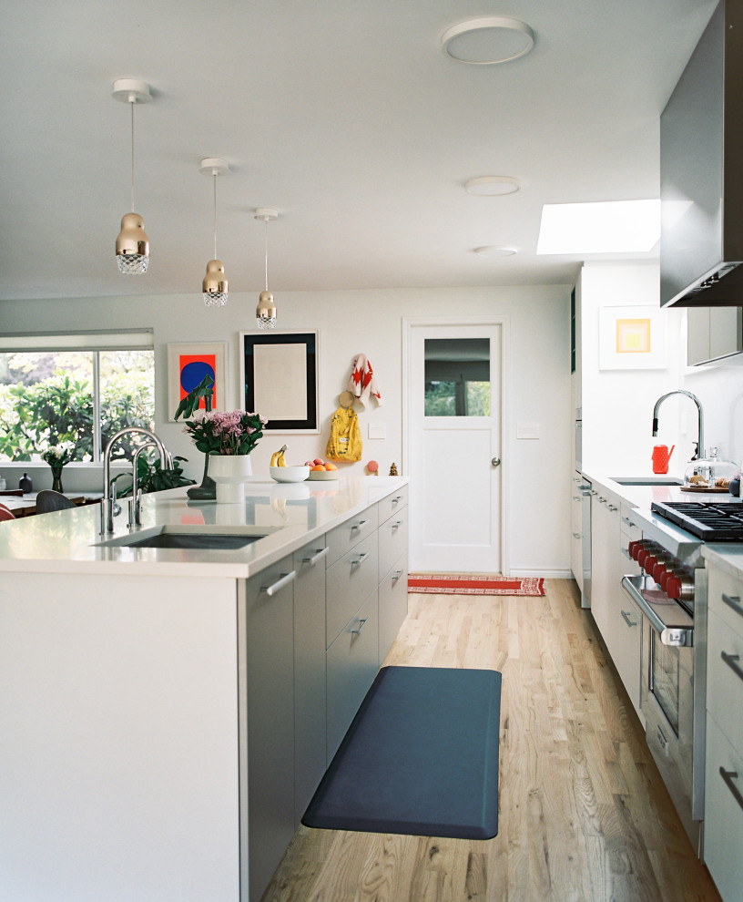 Foto på ett mellanstort funkis grå linjärt kök med öppen planlösning, med släta luckor, vita skåp, grått stänkskydd, stänkskydd i sten, rostfria vitvaror, ljust trägolv, en köksö och brunt golv