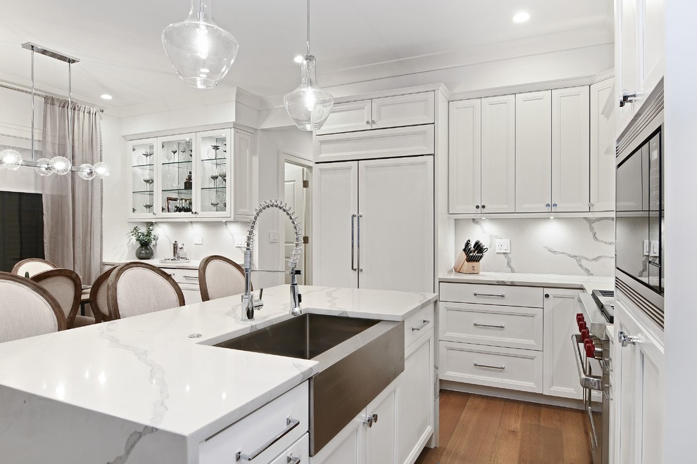 Photo of a traditional l-shaped kitchen/diner in Chicago with a belfast sink, recessed-panel cabinets, white cabinets, white splashback, stone slab splashback, integrated appliances, medium hardwood flooring, an island, brown floors and white worktops.