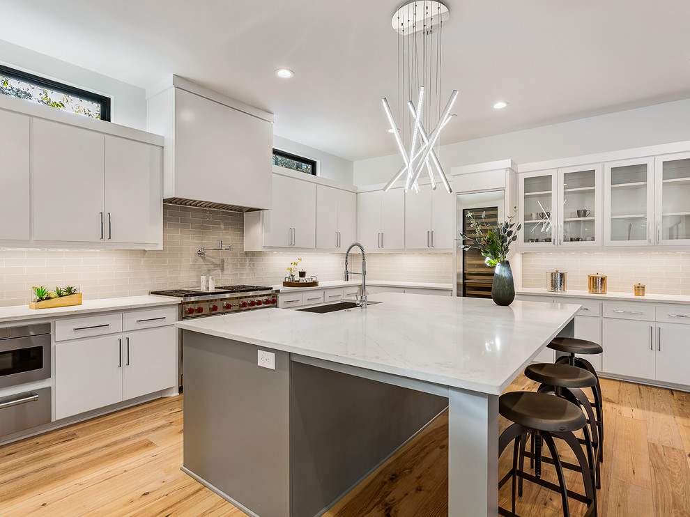 Example of a large trendy l-shaped light wood floor kitchen design in Austin with a farmhouse sink, flat-panel cabinets, white cabinets, quartz countertops, gray backsplash, stainless steel appliances, an island, white countertops and subway tile backsplash