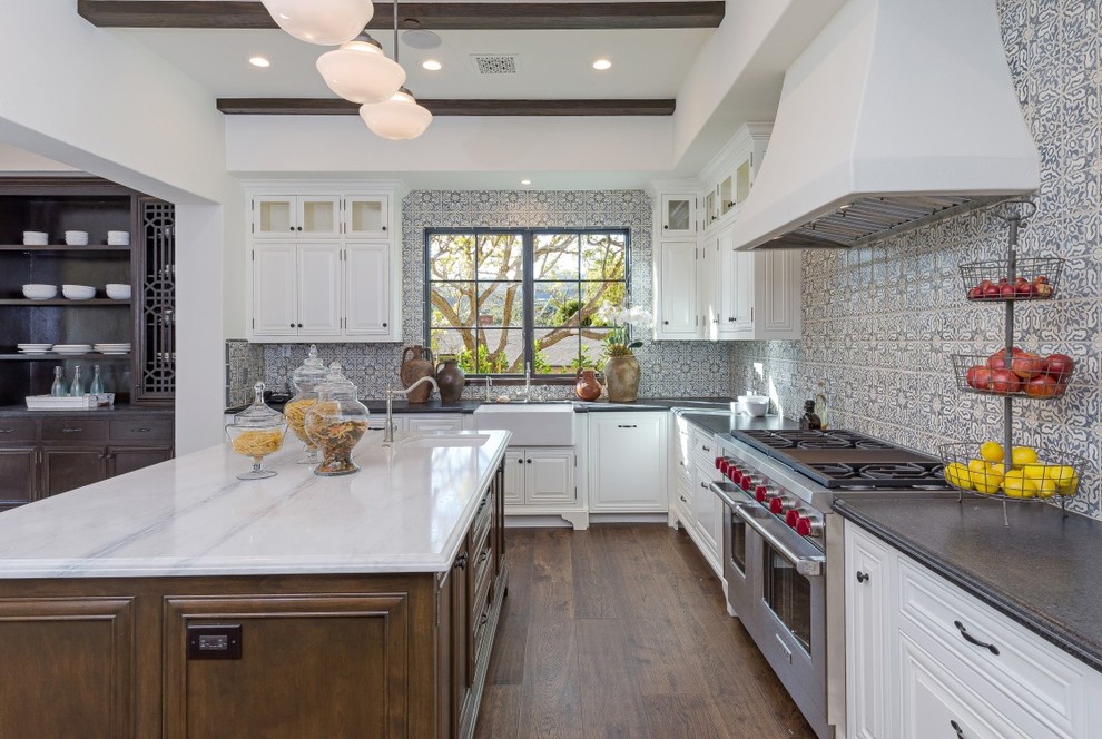 This is an example of a classic l-shaped kitchen in Los Angeles with a belfast sink, raised-panel cabinets, white cabinets, multi-coloured splashback, stainless steel appliances, dark hardwood flooring and an island.
