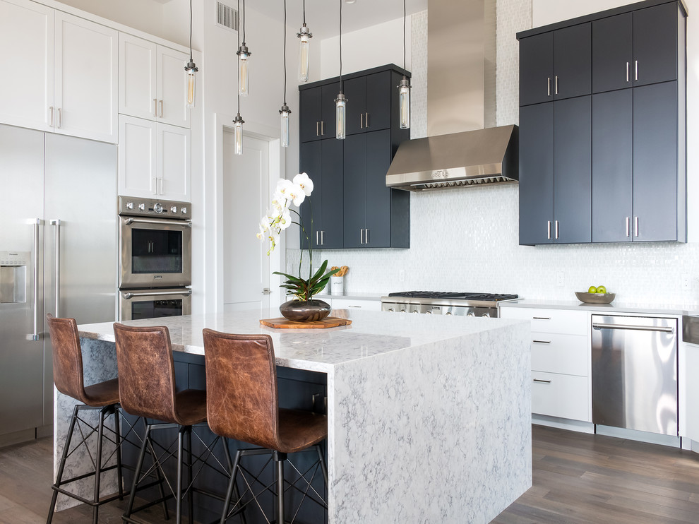 This is an example of a contemporary l-shaped kitchen in Austin with flat-panel cabinets, black cabinets, white splashback, stainless steel appliances, light hardwood flooring, an island, brown floors and grey worktops.