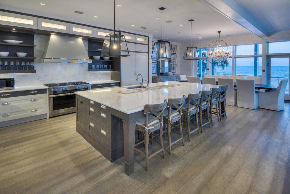 Example of a large beach style medium tone wood floor open concept kitchen design in Miami with an undermount sink, recessed-panel cabinets, white cabinets, marble countertops, white backsplash, stone tile backsplash, stainless steel appliances and an island
