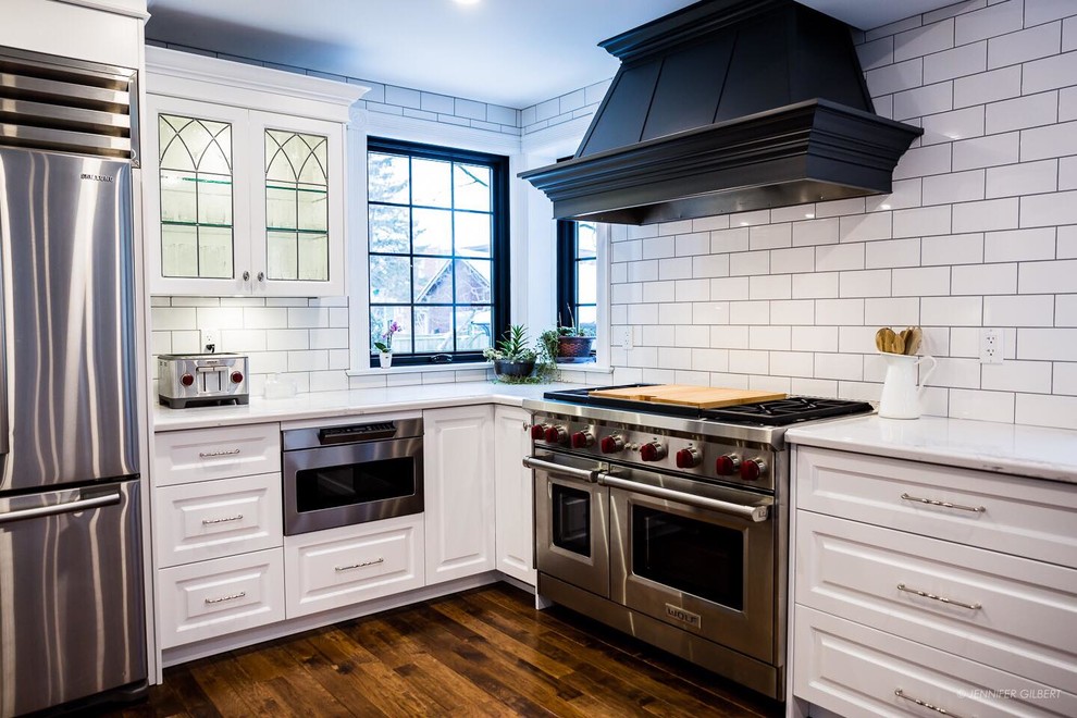 Inspiration for a large classic u-shaped kitchen/diner in Toronto with a belfast sink, raised-panel cabinets, white cabinets, engineered stone countertops, white splashback, ceramic splashback, stainless steel appliances, medium hardwood flooring, a breakfast bar and brown floors.