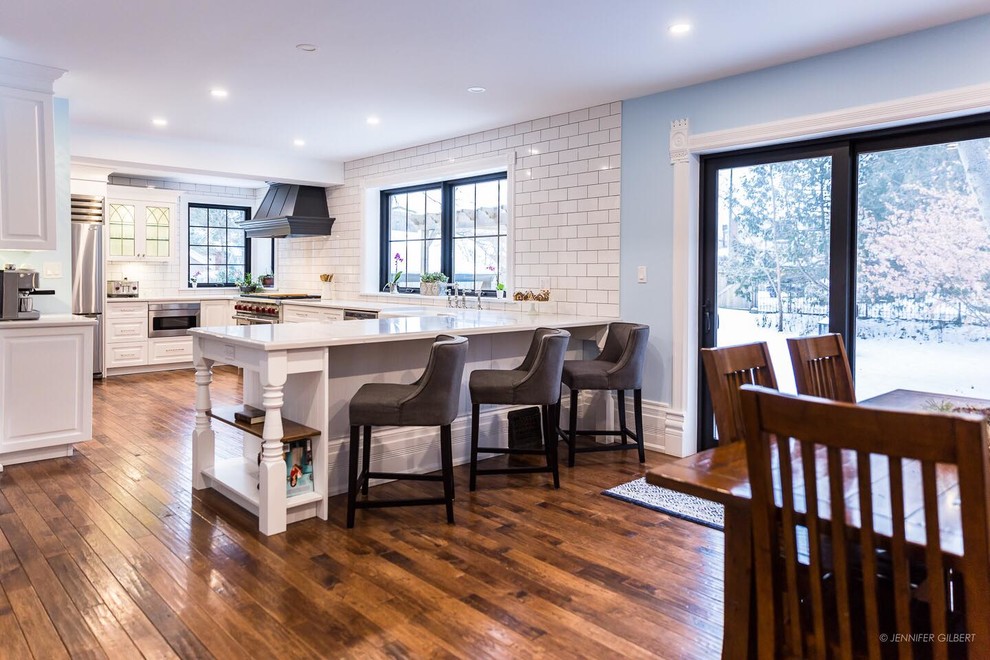 Large elegant u-shaped medium tone wood floor and brown floor eat-in kitchen photo in Toronto with a farmhouse sink, raised-panel cabinets, white cabinets, quartz countertops, white backsplash, ceramic backsplash, stainless steel appliances and a peninsula
