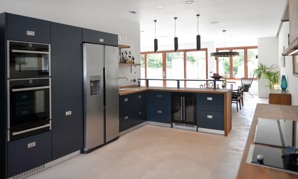 Modern inredning av ett mellanstort kök med öppen planlösning, med en nedsänkt diskho, släta luckor, blå skåp, träbänkskiva, svart stänkskydd, stänkskydd i keramik, svarta vitvaror, betonggolv och en köksö
