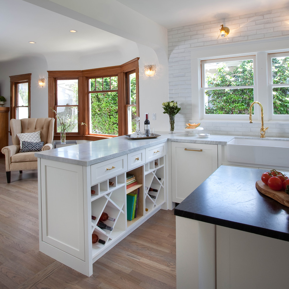 Example of a transitional u-shaped medium tone wood floor eat-in kitchen design in San Diego with a farmhouse sink, shaker cabinets, white cabinets, soapstone countertops, white backsplash and an island