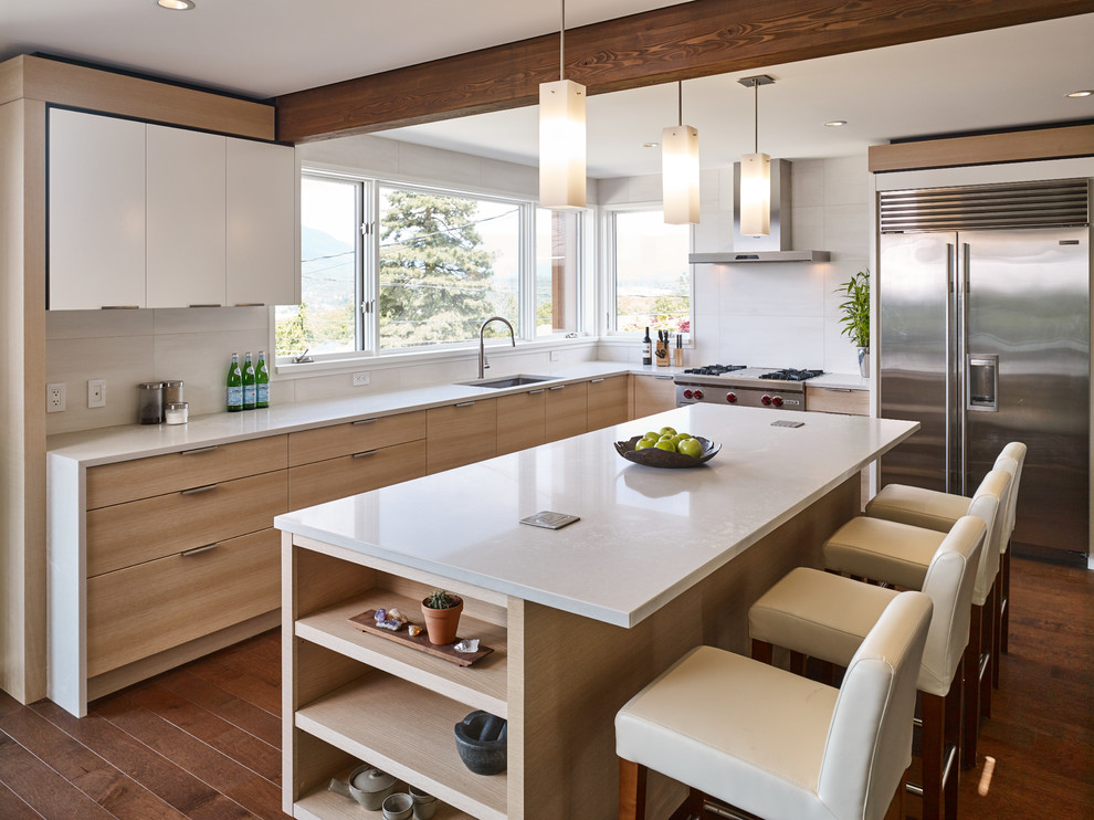 Trendy l-shaped dark wood floor and brown floor kitchen photo in Vancouver with an undermount sink, flat-panel cabinets, light wood cabinets, white backsplash, stainless steel appliances, an island and gray countertops