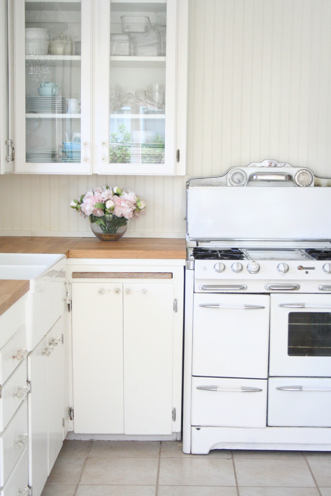 Aménagement d'une petite cuisine américaine romantique en L avec un évier de ferme, un placard à porte vitrée, des portes de placard blanches, un plan de travail en bois, une crédence blanche, un électroménager en acier inoxydable, un sol en carrelage de céramique et aucun îlot.