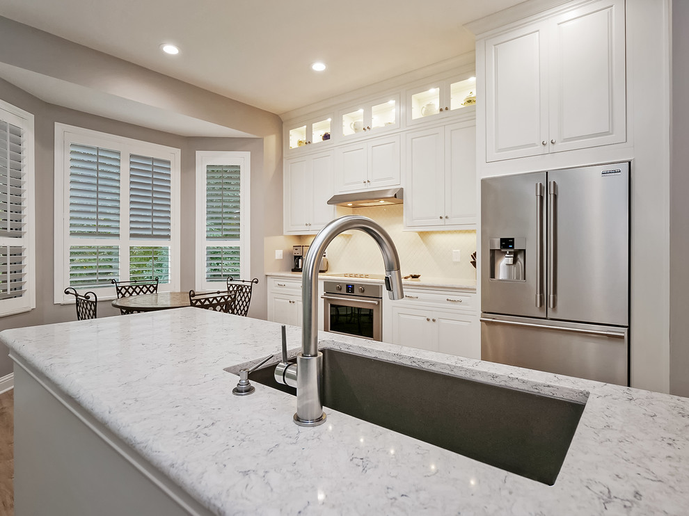 Klassisk inredning av ett mellanstort grå grått kök, med en undermonterad diskho, luckor med profilerade fronter, vita skåp, bänkskiva i kvarts, vitt stänkskydd, stänkskydd i keramik, rostfria vitvaror, vinylgolv, en halv köksö och beiget golv
