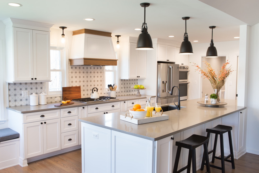 Klassisk inredning av ett stort grå grått kök, med en undermonterad diskho, vita skåp, bänkskiva i kvartsit, vitt stänkskydd, stänkskydd i keramik, rostfria vitvaror, mellanmörkt trägolv, brunt golv, en halv köksö och skåp i shakerstil