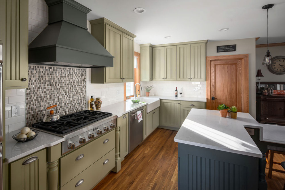 Large classic l-shaped kitchen/diner in Columbus with a belfast sink, recessed-panel cabinets, green cabinets, engineered stone countertops, white splashback, ceramic splashback, stainless steel appliances, medium hardwood flooring, multiple islands, brown floors and white worktops.