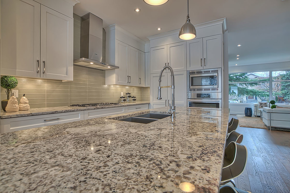 Mid-sized transitional l-shaped medium tone wood floor and brown floor eat-in kitchen photo in Calgary with a double-bowl sink, shaker cabinets, white cabinets, granite countertops, gray backsplash, ceramic backsplash, stainless steel appliances and an island