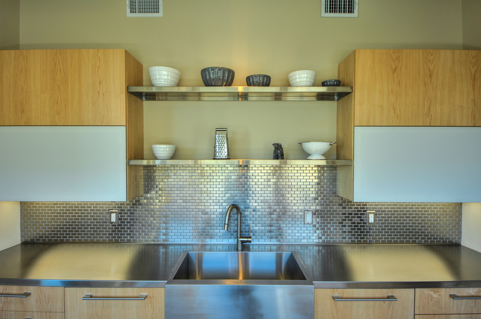 Cette image montre une cuisine design en bois clair avec un évier de ferme, un placard à porte plane, un plan de travail en inox et une crédence métallisée.