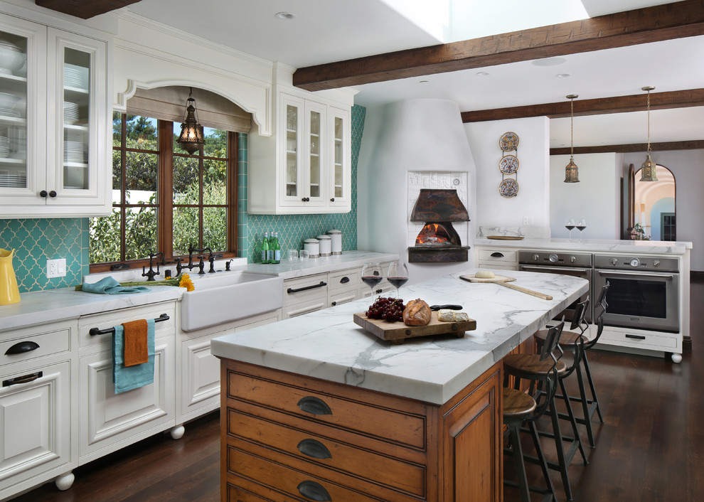 Inredning av ett medelhavsstil kök, med en rustik diskho, luckor med upphöjd panel, vita skåp, blått stänkskydd, rostfria vitvaror, mörkt trägolv och en köksö