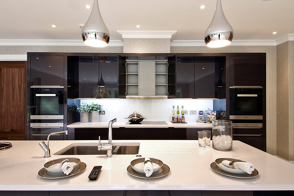 Große Moderne Wohnküche in L-Form mit Doppelwaschbecken, flächenbündigen Schrankfronten, grauen Schränken, Quarzwerkstein-Arbeitsplatte, Küchenrückwand in Weiß, Glasrückwand, schwarzen Elektrogeräten und Kücheninsel in Buckinghamshire