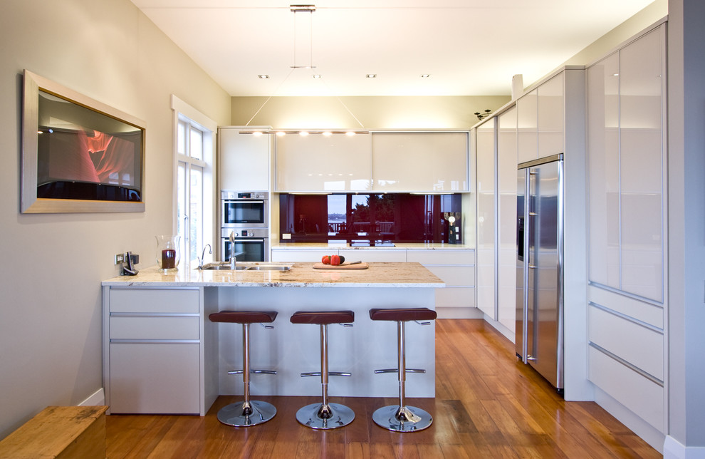 Medium sized contemporary u-shaped kitchen/diner in Auckland with a submerged sink, glass-front cabinets, red splashback, glass sheet splashback, stainless steel appliances, medium hardwood flooring and a breakfast bar.