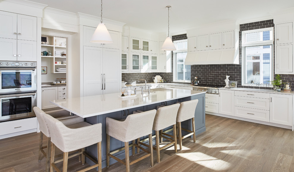 Large nautical l-shaped open plan kitchen in Grand Rapids with stainless steel appliances, shaker cabinets, white cabinets, brown splashback, metro tiled splashback, medium hardwood flooring and an island.