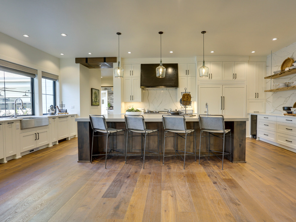 Photo of an expansive country u-shaped open plan kitchen in Portland with a belfast sink, shaker cabinets, white cabinets, engineered stone countertops, white splashback, stone slab splashback, integrated appliances, medium hardwood flooring, an island and brown floors.