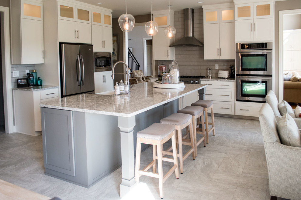 Photo of a large romantic l-shaped kitchen/diner in Louisville with a belfast sink, shaker cabinets, white cabinets, engineered stone countertops, grey splashback, glass tiled splashback, stainless steel appliances, ceramic flooring, an island, grey floors and white worktops.