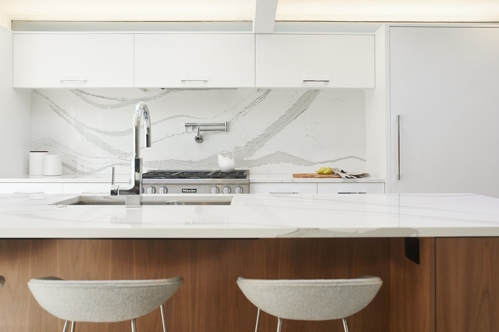 グランドラピッズにある高級な巨大なミッドセンチュリースタイルのおしゃれなキッチン (アンダーカウンターシンク、フラットパネル扉のキャビネット、白いキャビネット、コンクリートカウンター、白いキッチンパネル、石スラブのキッチンパネル、パネルと同色の調理設備、淡色無垢フローリング、茶色い床、白いキッチンカウンター) の写真